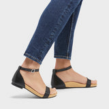  Person in jeans wearing black leather sandal with a padded heel cup and toe strap. The footbed is nude, and fitted with arch support. 